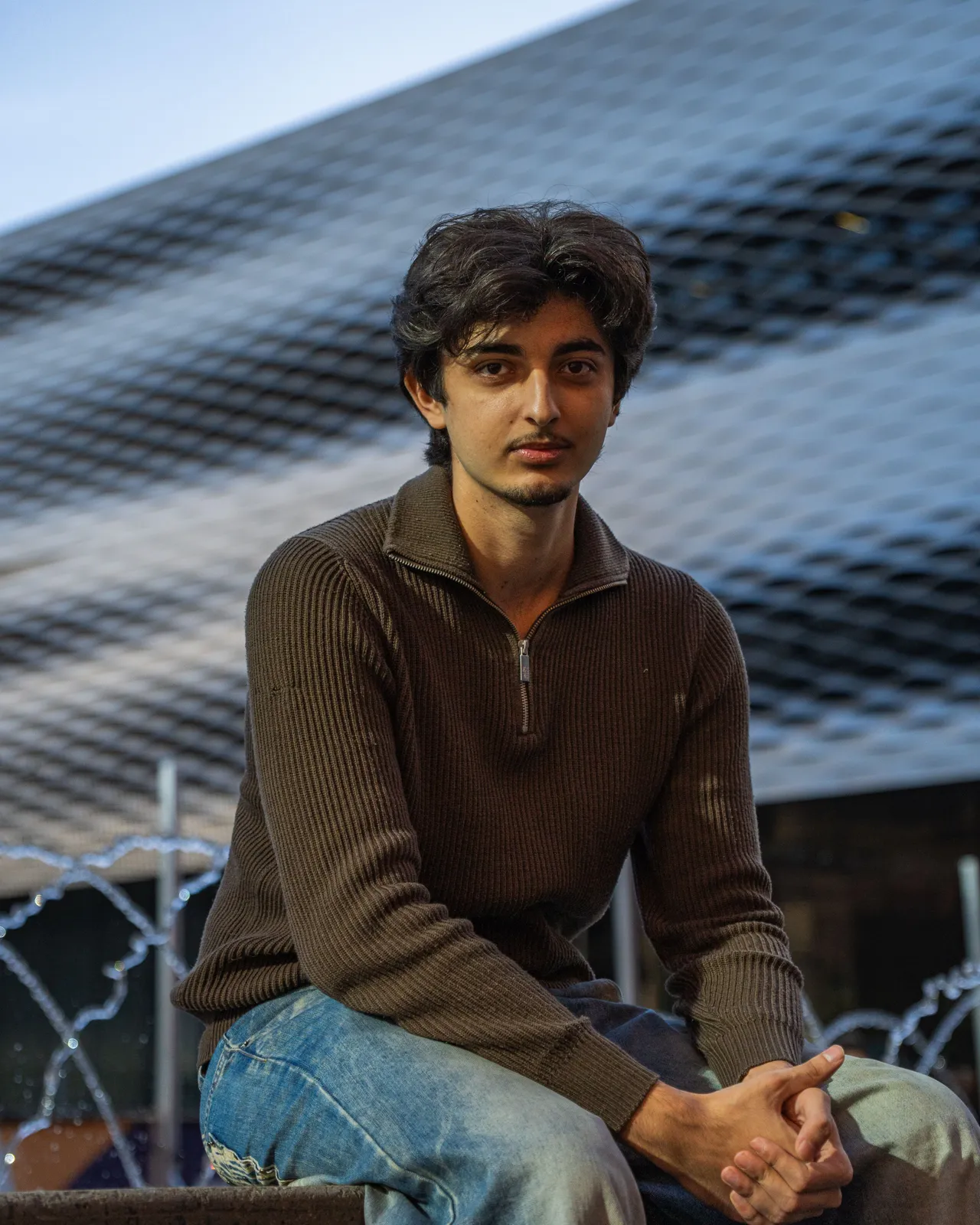 Aryan sitzt auf einer Mauer vor einer geschwungenen Metallfassade.