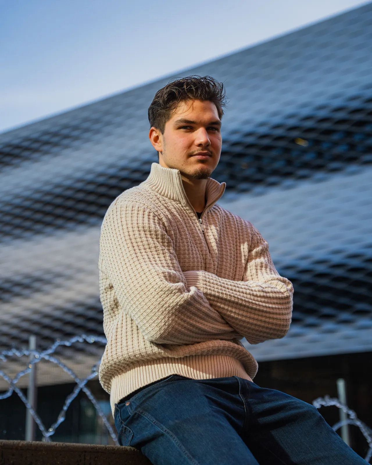 Moritz sitzt mit verschränkten Armen auf einem Brunnenrand vor einer geschwungenen Metallfassade.